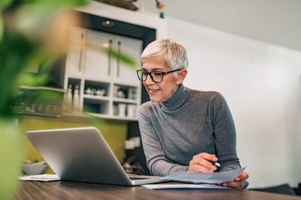 A woman sitting at her kitchen table and looking at a laptop while filling out paperwork.