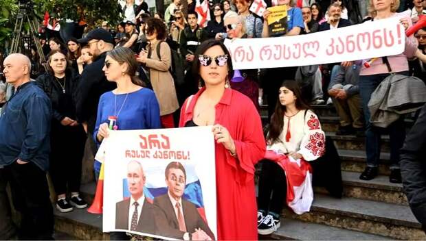 Демонстрант в Тбилиси несет табличку с портретами Владимира Путина и Бидзины Иванишвили под надписью: «Нет российскому закону».  (ВАНО ШЛАМОВ / AFP)