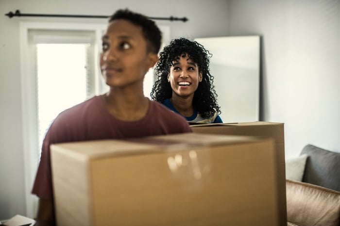 A couple smiles as they enter their new house carrying moving boxes.