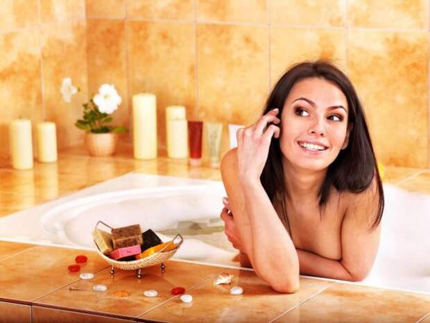 Симпатичная девушка улыбается, сидя в ванной, слева от неё вазочка с мылом, на заднем плане свечи и цветок в горшке