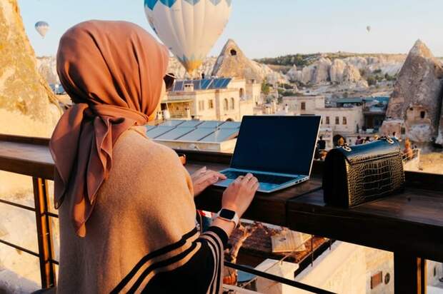 A person sitting on a balcony and typing on a laptop while looking over a scenic view filled with stony hills, low buildings, and hot air balloons.