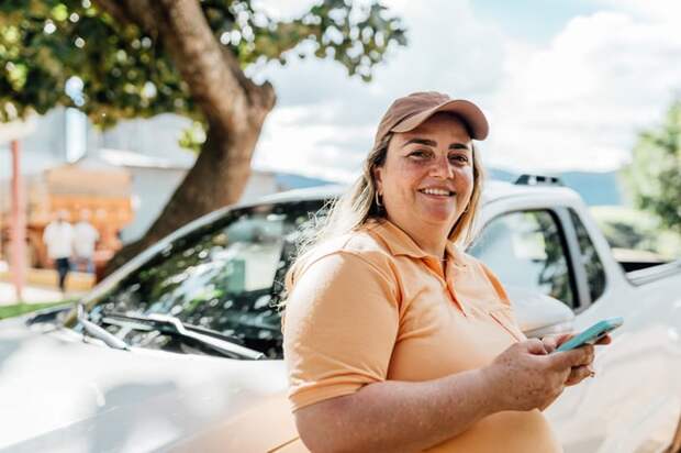 Person in a polo shirt and ballcap, leaning on a car and smiling. 