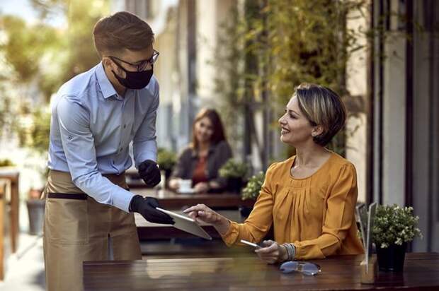 A waiter wearing a medical mask and gloves while taking the order of a woman seated at an outdoor table at a restaurant.