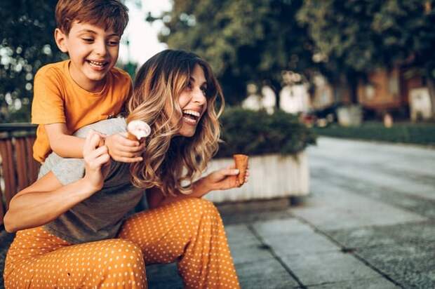 A mom and her son laughing and holding ice cream cones while he climbs on her back from a bench under a tree.