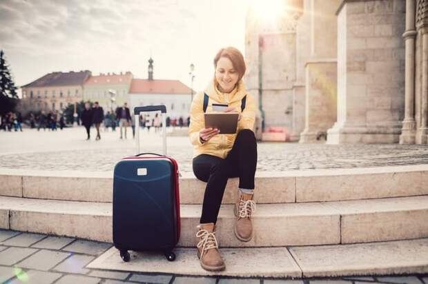 A woman sitting in a sunny plaza next to her suitcase while she looks something up on her tablet and holds her credit card.