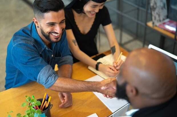 Two people meeting with a third person in an office and shaking hands across the desk.