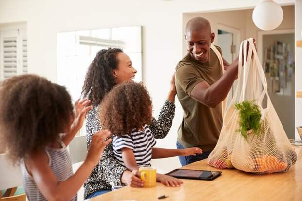 A father bringing home bags of groceries to his family waiting in the kitchen.