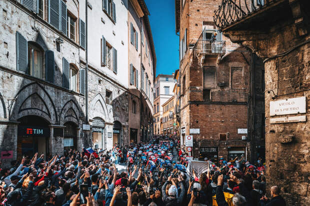 Зрители на узких улочках Сиены, Италия, наблюдают за прохождением группы велосипедистов