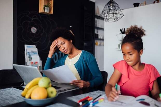 A mom looking worried while reviewing papers at the kitchen table next to her young daughter doing homework.