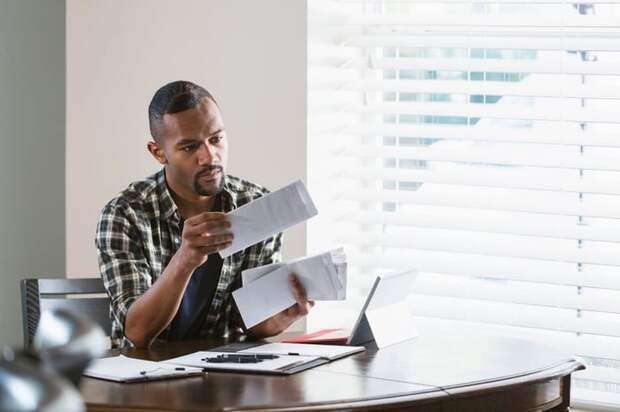 A person with facial hair sorts through bills at the kitchen table with a neutral expression.