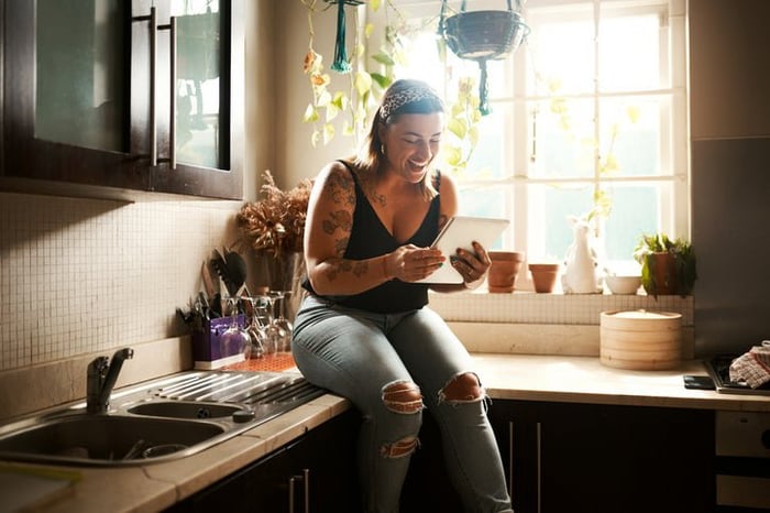 A woman sitting on the kitchen counter in her apartment and smiling at a tablet she's holding.