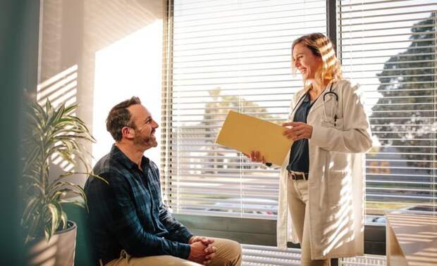 A doctor standing in front of a sunny window in her office and speaking with her male patient sitting in a chair.