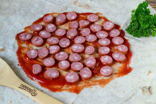 На рабочей поверхности расстелите большой лист лаваша, смажьте кетчупом. Выложите первым слоем колбасу.