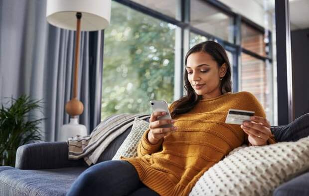 A woman sitting comfortably on her couch looking at her phone and holding a credit card.