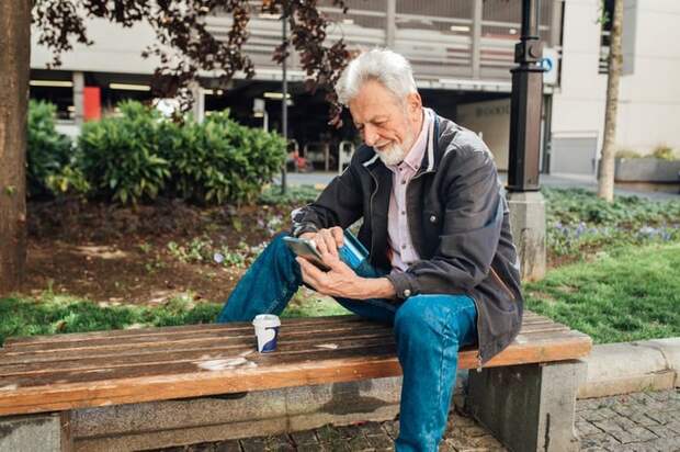 An older gentleman sits on a park bench, looking at his phone while also holding his credit card.