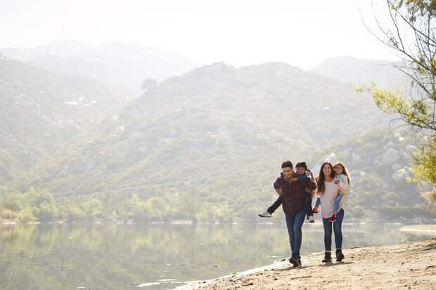 Two young kids piggybacking on their parents as they hike next to a lake with hills in the background.