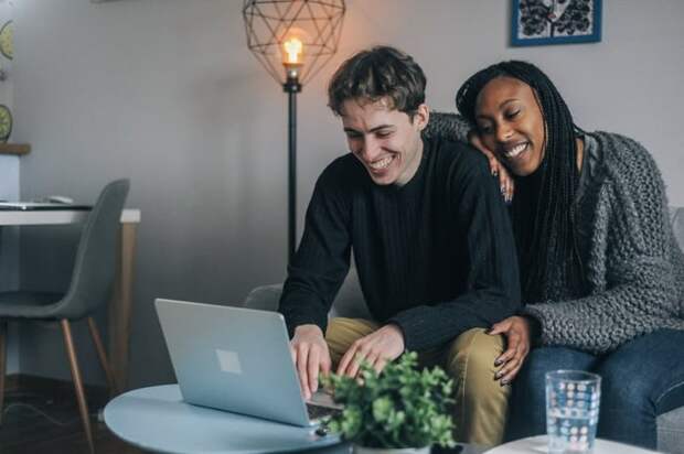A smiling couple sitting on the couch while looking something up on their laptop.