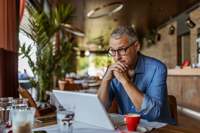 A man sitting in front of his laptop at a cafe table and looking stressed.