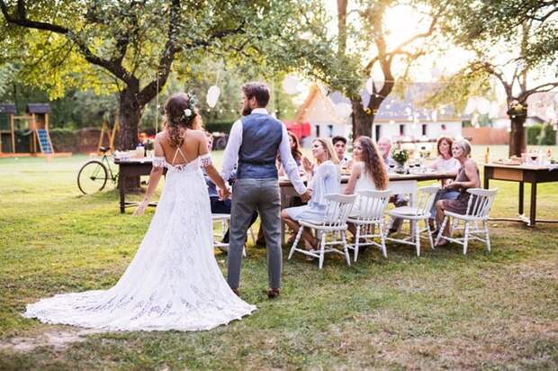 A bride and groom holding hands and walking toward their guests seated at a table in a decorated backyard.