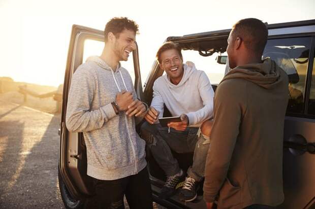 Three smiling men standing around their car and planning their road trip route on a phone.