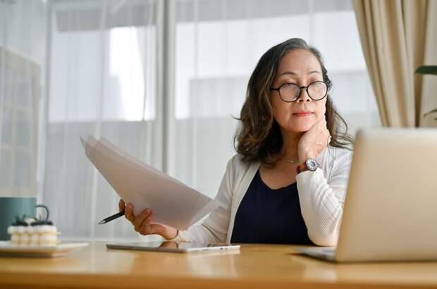 A businessperson holding paperwork in their right hand while looking at an open laptop in their office.